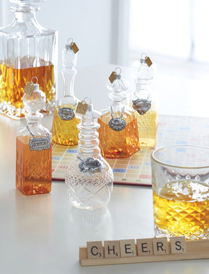 Image of five Christmas ornaments that are  clear glass alcohol decanters with translucent painted liquor inside. Each decanter has a label that includes whisky, bourbon, vodka, tequila, and scotch
