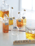Image of five Christmas ornaments that are  clear glass alcohol decanters with translucent painted liquor inside. Each decanter has a label that includes whisky, bourbon, vodka, tequila, and scotch